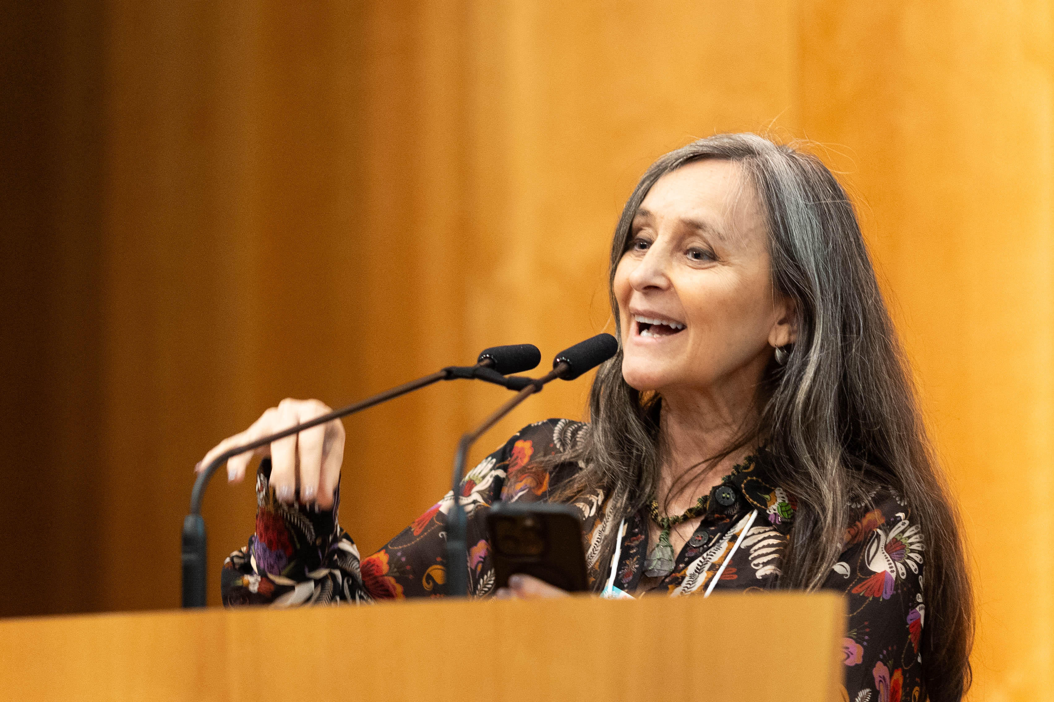A Prof.ª Viviana Martinez-Bianchi fala em um púlpito de madeira, segurando o telefone, com dois microfones à sua frente.